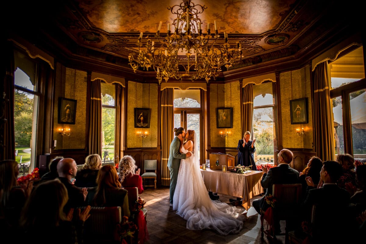 Bruiloftsceremonie koppel zoent in elegante, sierlijke kamer met houten lambrisering, kroonluchters, hoge ramen, gasten klappen.