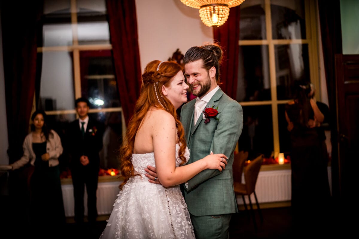 Bruiloftsdans: bruid in witte jurk en bruidegom in lichtgroen pak dansen samen, omringd door gasten en rode gordijnen.