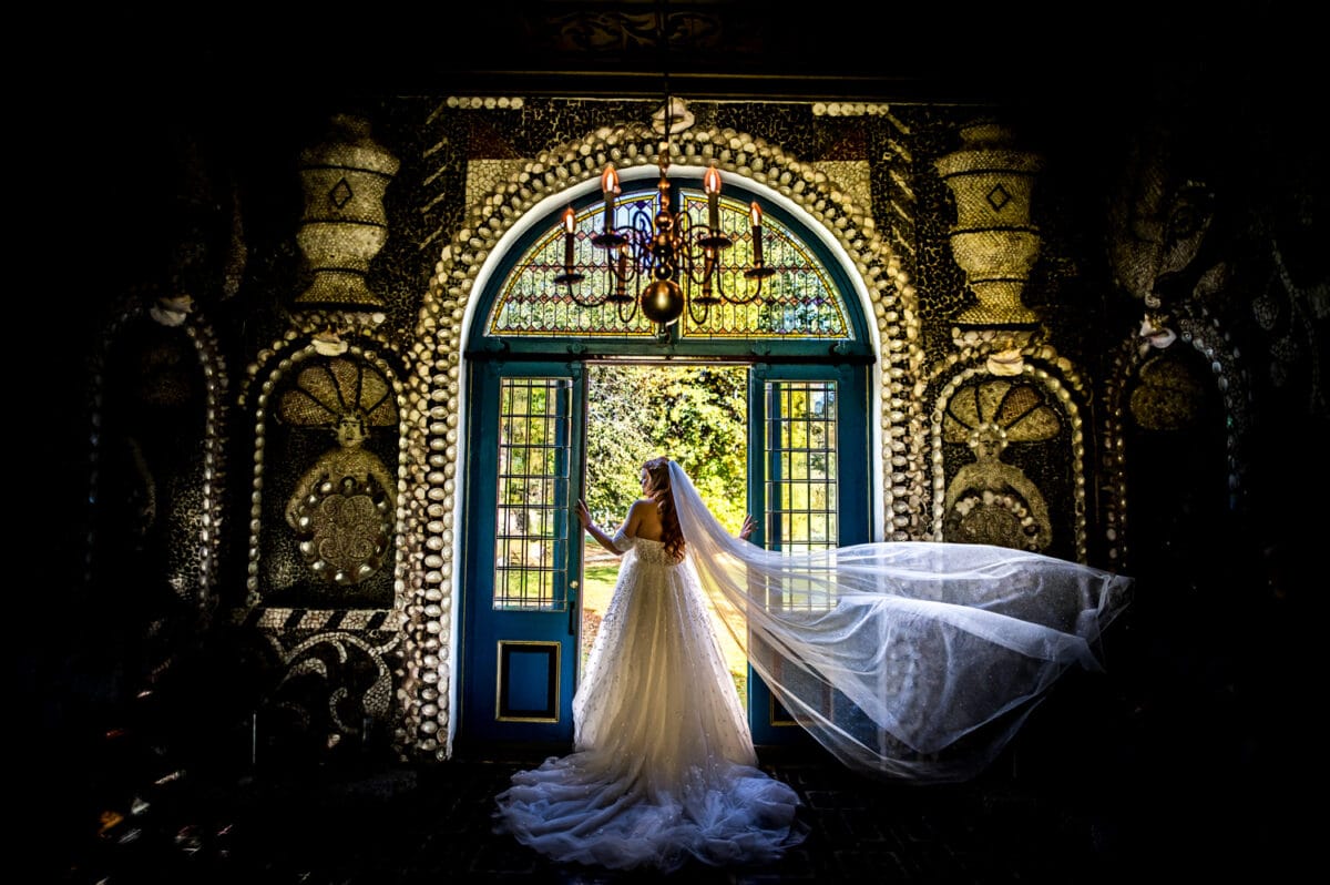 Bruid in witte jurk en sluier staat voor sierlijke blauwe dubbele deuren met mozaïektegels, zonlicht stroomt, kroonluchter erboven.