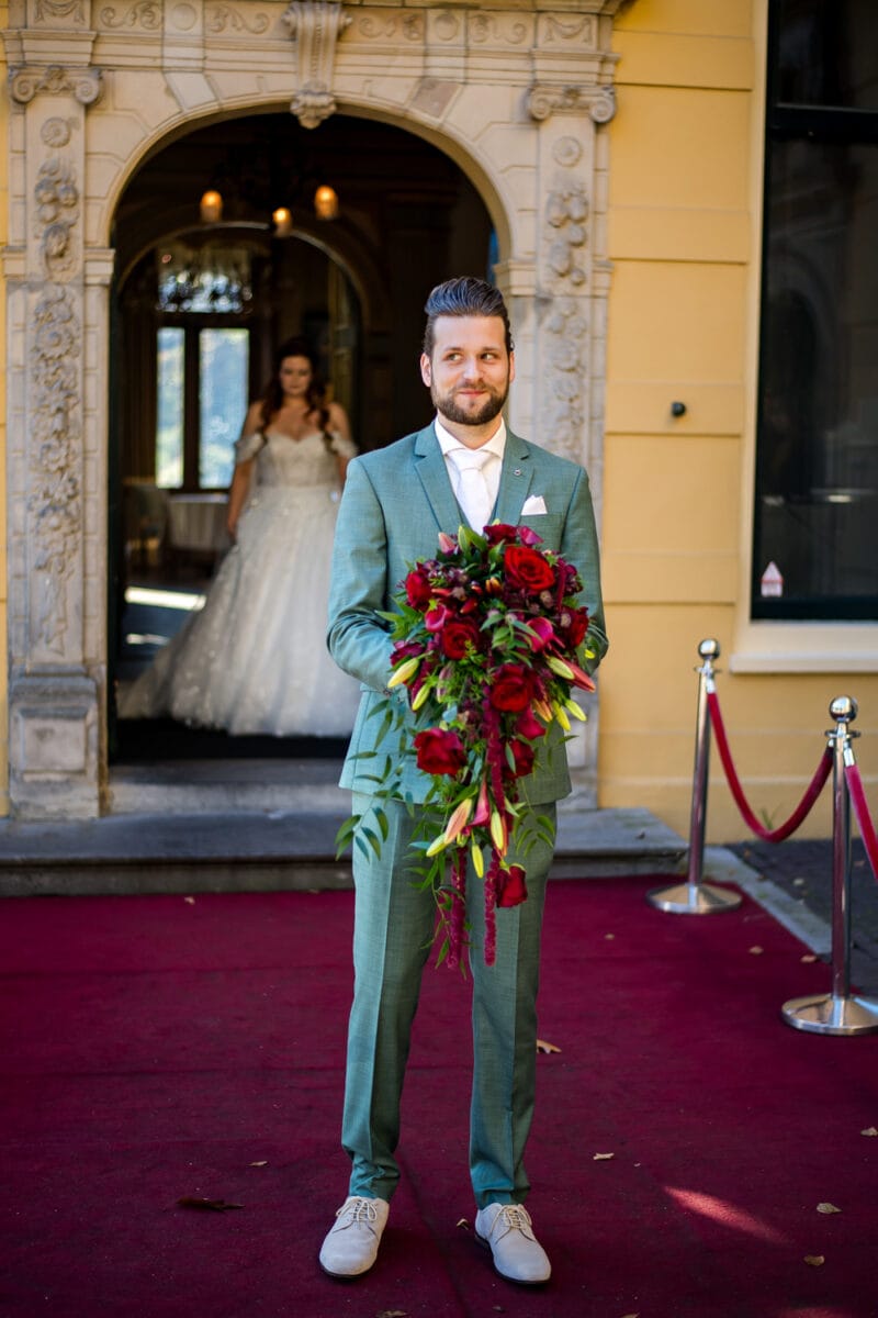 Bruidegom in lichtgroen pak op rode loper met boeket rozen en lelies, bruid in witte jurk op achtergrond in sierlijke zaal.