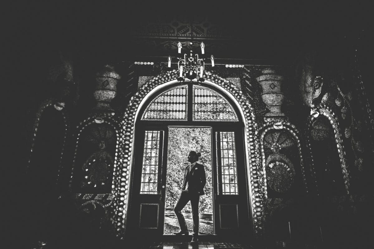 Silhouet van persoon in formele kleding voor grote sierdeuren en boog, met kroonluchter en fel licht, luxe interieur.
