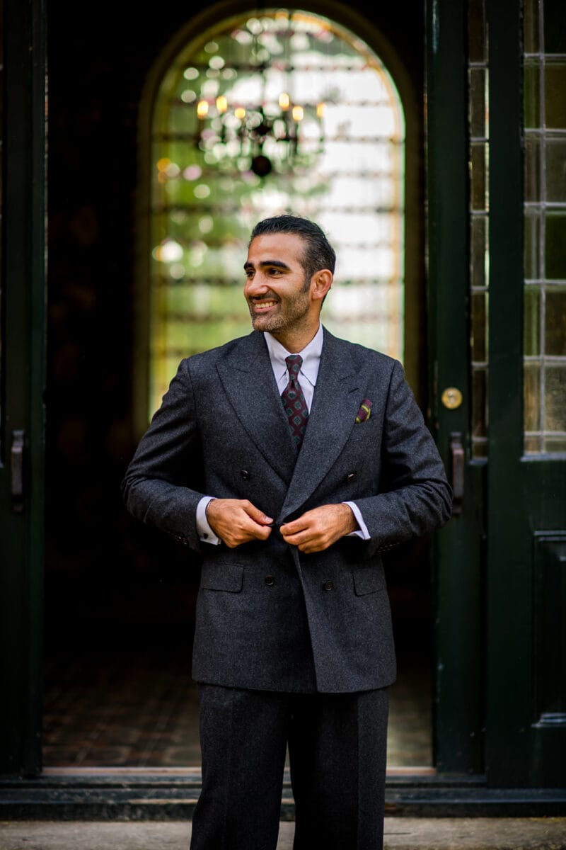 Man in houtskool pak met dubbele rij knopen en stropdas glimlacht, knoopt jasje dicht bij groene deuren met boograam en kroonluchter.