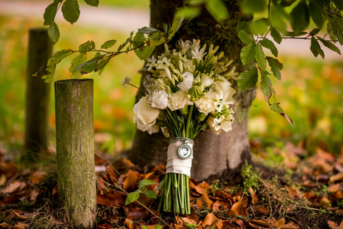 Bruidsboeket witte bloemen met pluis en foto-hanger bij boomstam, omringd door groene bladeren en gras buiten, bruin blad op de grond.