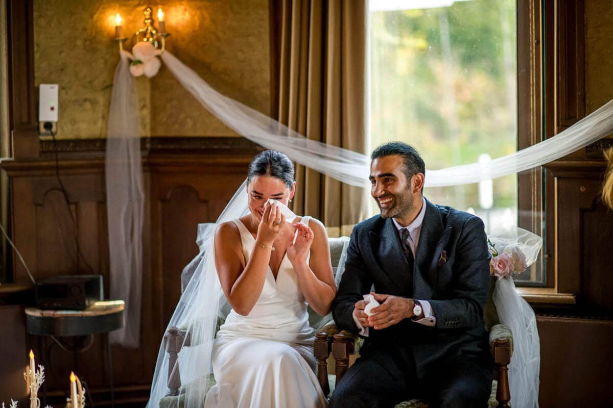 Emotionele bruid in witte jurk veegt tranen weg naast lachende bruidegom in zwart pak, bij sfeervolle bruiloft binnen.