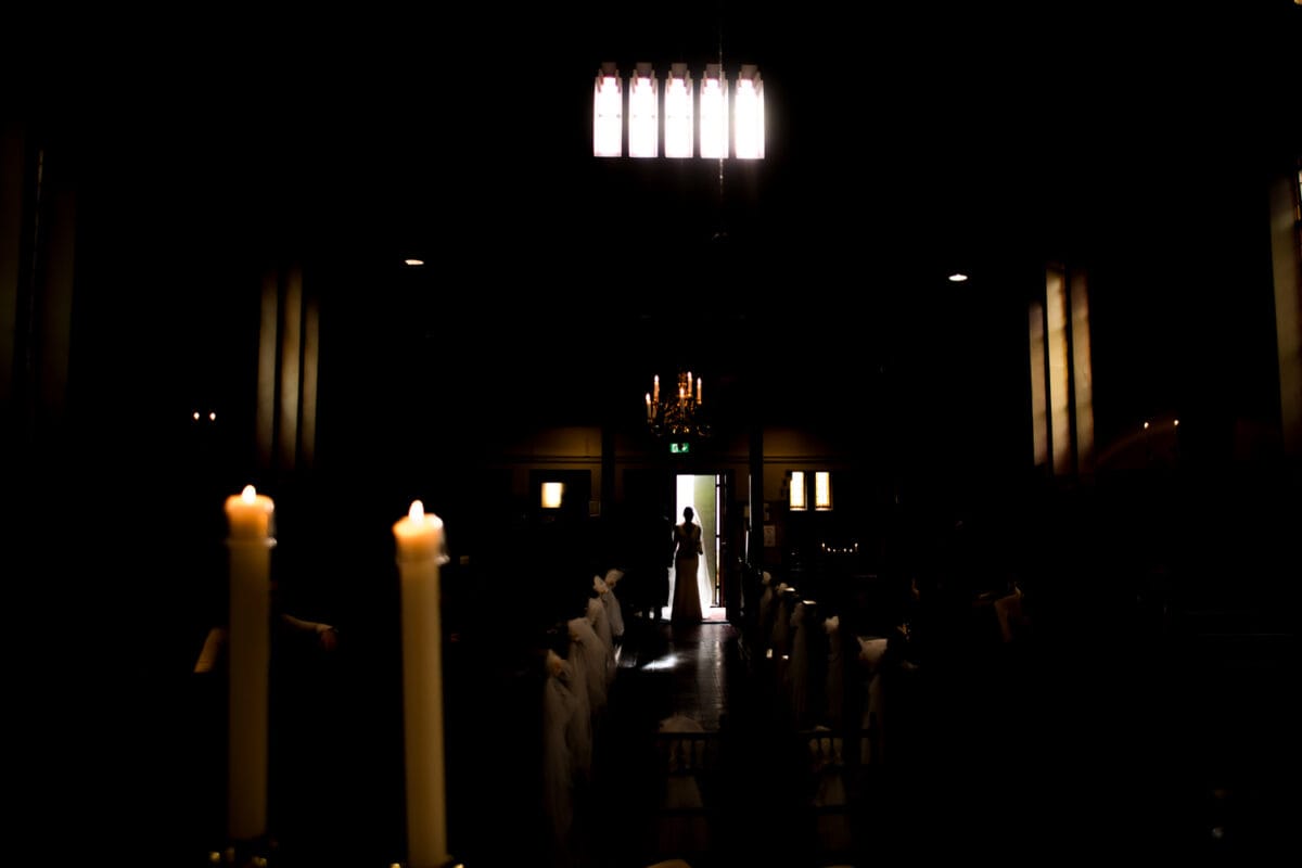 Kerkinterieur met gloeiende kaarsen langs het gangpad, silhouet van de bruid bij de heldere ingang, dramatisch contrast van licht en schaduw.