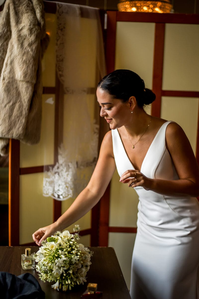 Vrouw in witte jurk met V-hals rangschikt witte bloemen op tafel, kanten sluier en bontjas hangen aan houten achterwand.