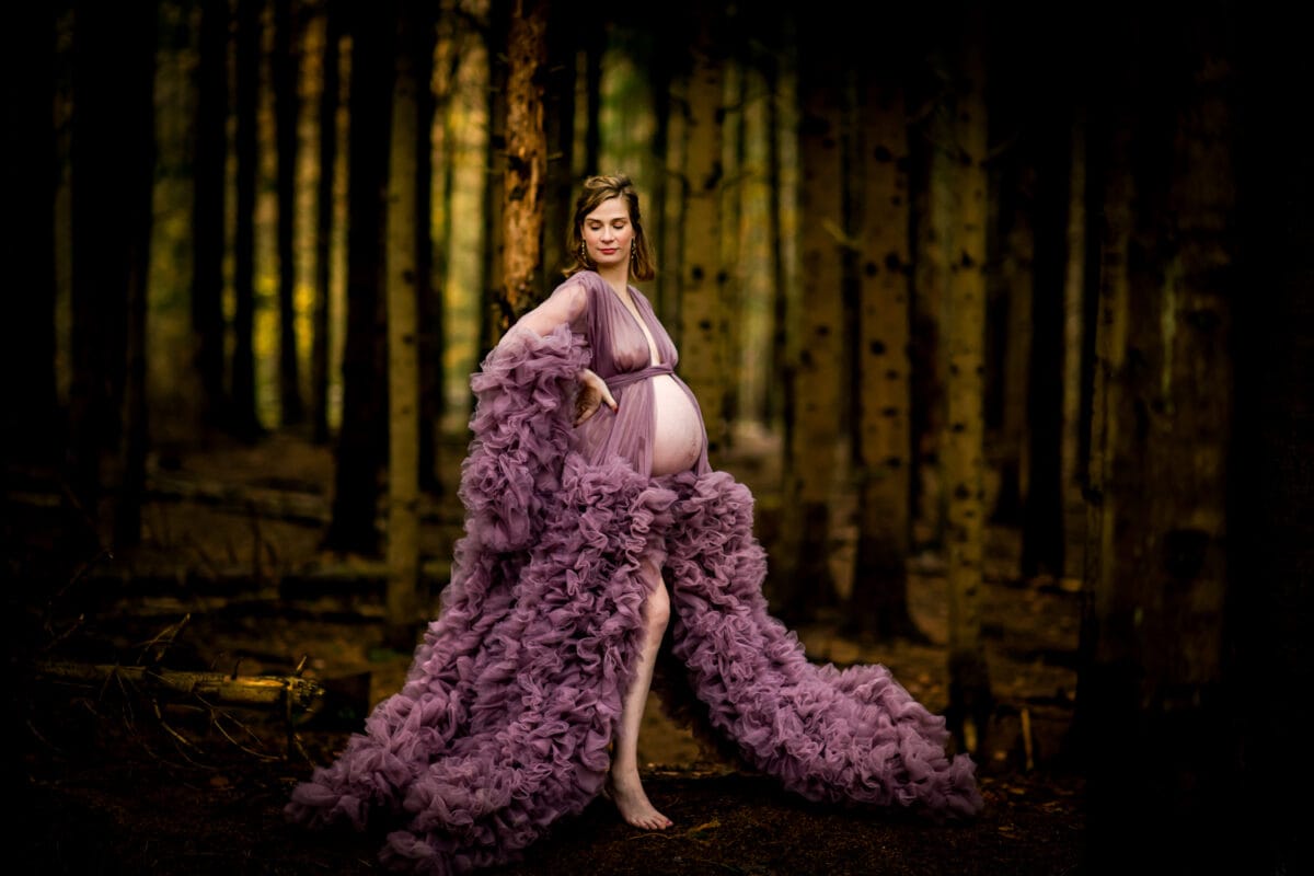Zwangere vrouw in vloeiende lavendeljurk staat blootsvoets in schemerig bos, houdt buik vast, omringd door hoge bomen en zacht gedimd licht.