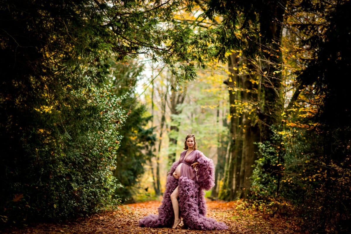 Zwangere vrouw in mauve jurk staat op herfstig bospad, buik wiegend; levendig gebladerte en hoge bomen creëren serene scène.