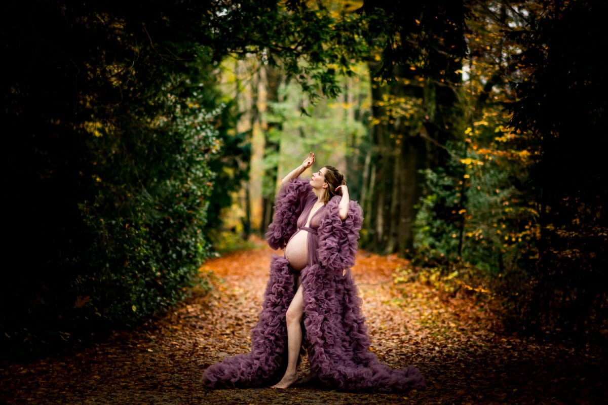 Zwangere vrouw in paarse jurk met ruches op herfstig bospad, armen omhoog, natuurlijk licht, zwangerschapsfotoshoot.