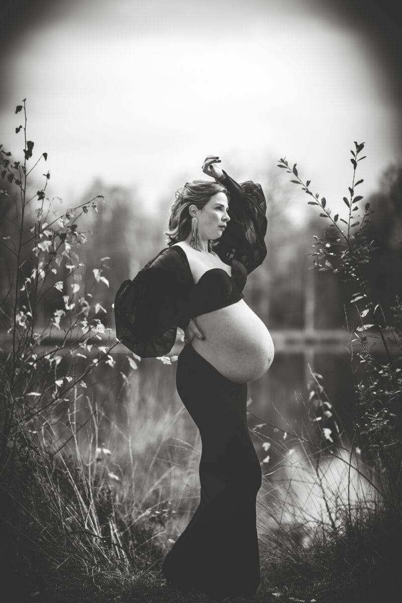 Zwangerschapsfotoshoot bij het water: vrouw in zwarte jurk, handen op buik, zwart-wit, omgeven door gras en bladeren.