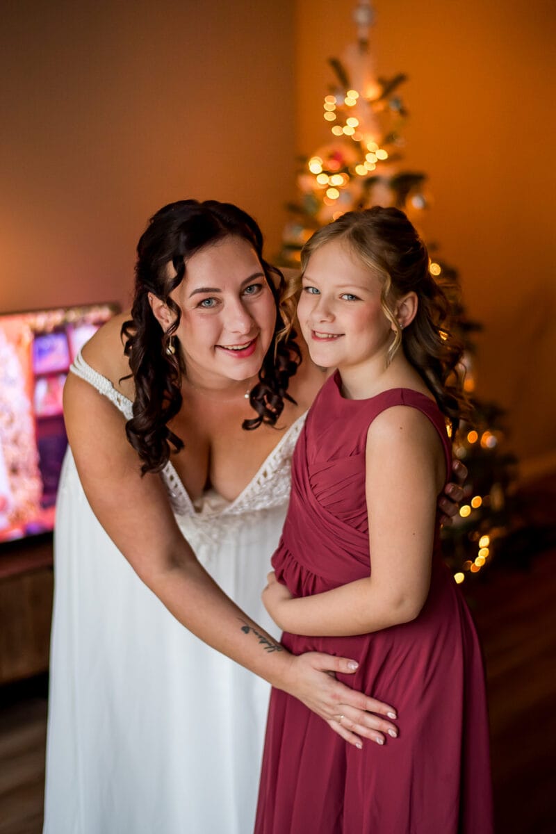 Vrouw in witte jurk en meisje in bordeaux jurk lachen voor versierde kerstboom in warm verlichte kamer, kerst sfeer.