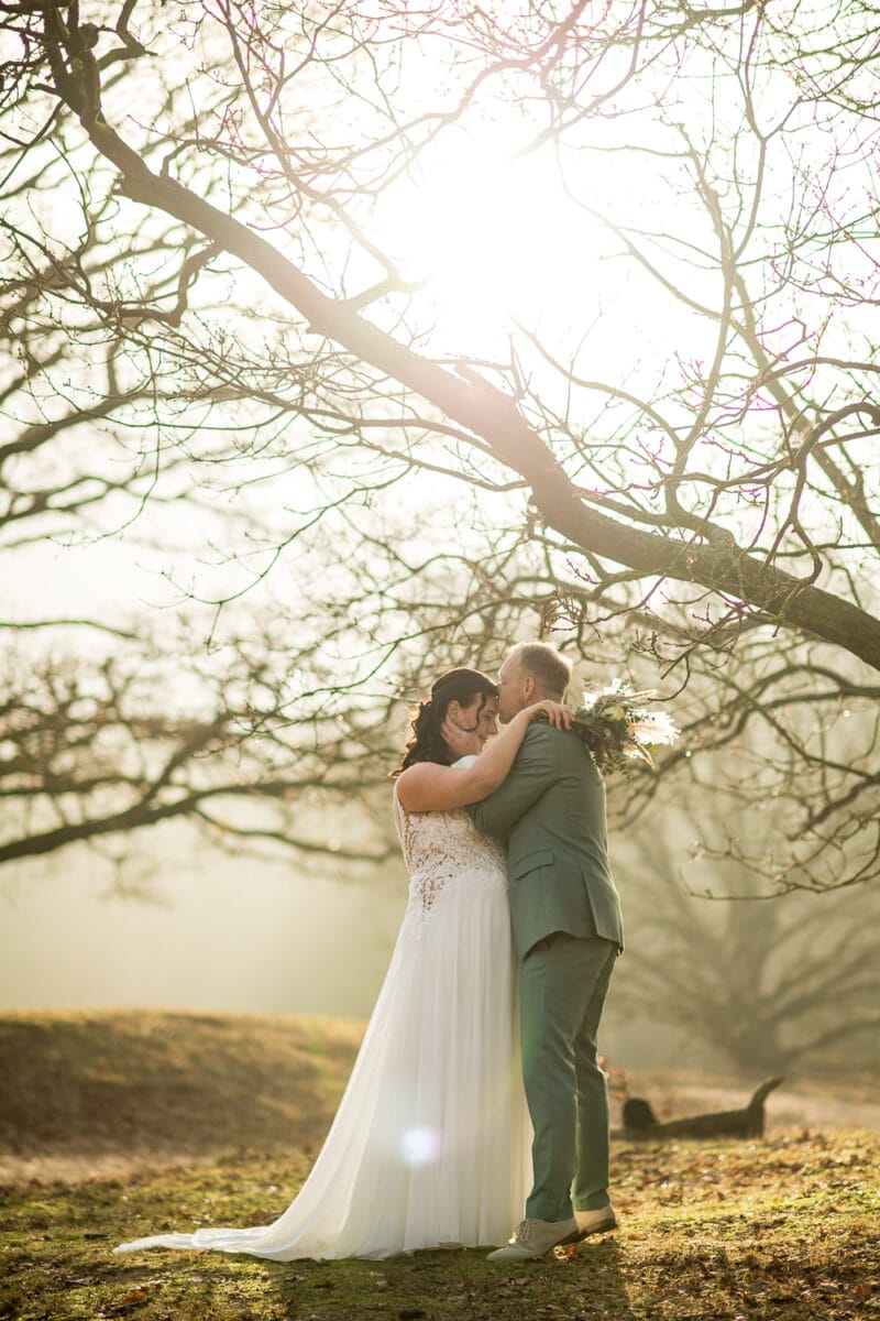 Romantische bruiloftsfoto buiten: bruid in witte jurk en bruidegom in lichtgrijs pak kussen bij zonsondergang onder boerenkool takken.