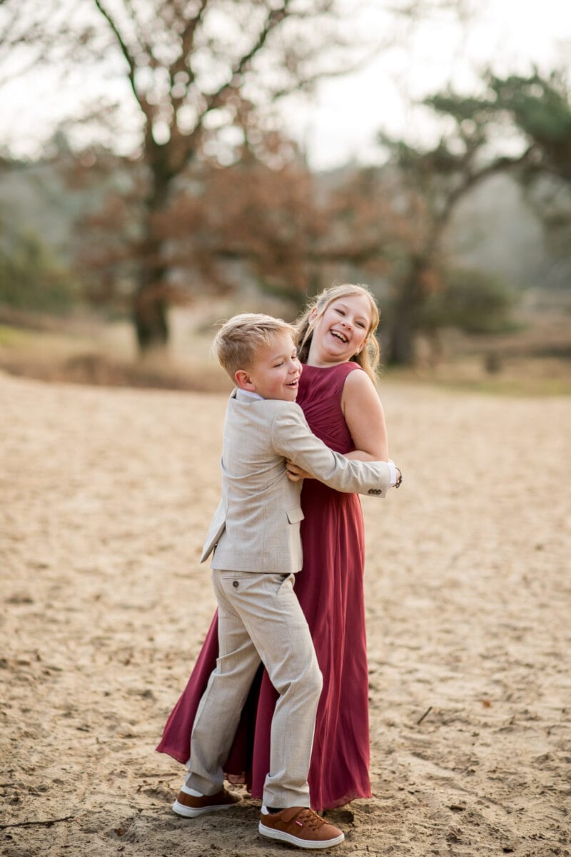Jongen in grijs pak en meisje in bordeaux jurk omhelzen elkaar lachend buiten op zand, bruine schoenen, groene bomen vaag op achtergrond.