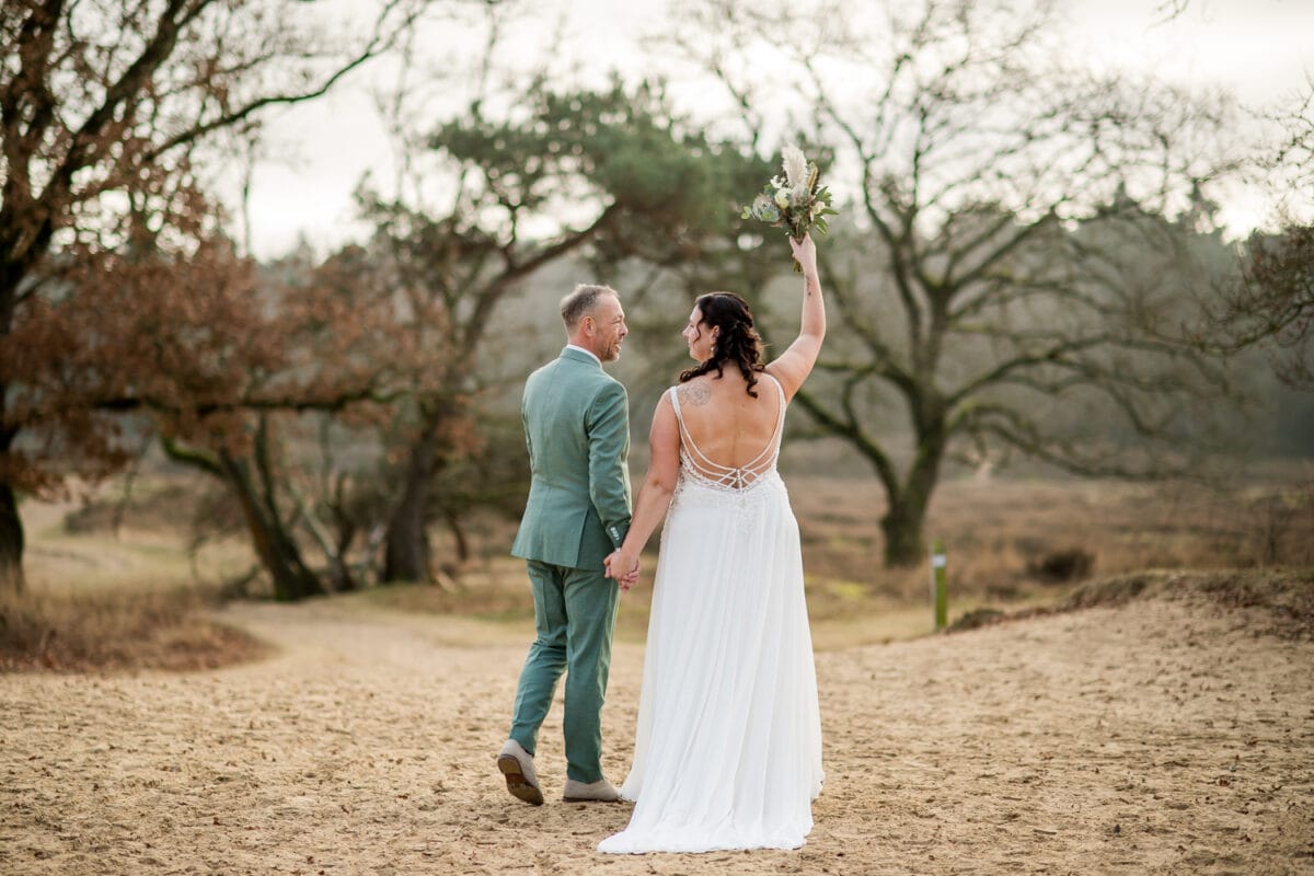 Bruid in witte jurk en bruidegom in lichtgroen pak lopen hand in hand over zandpad, boeket omhoog, buiten bruiloft.