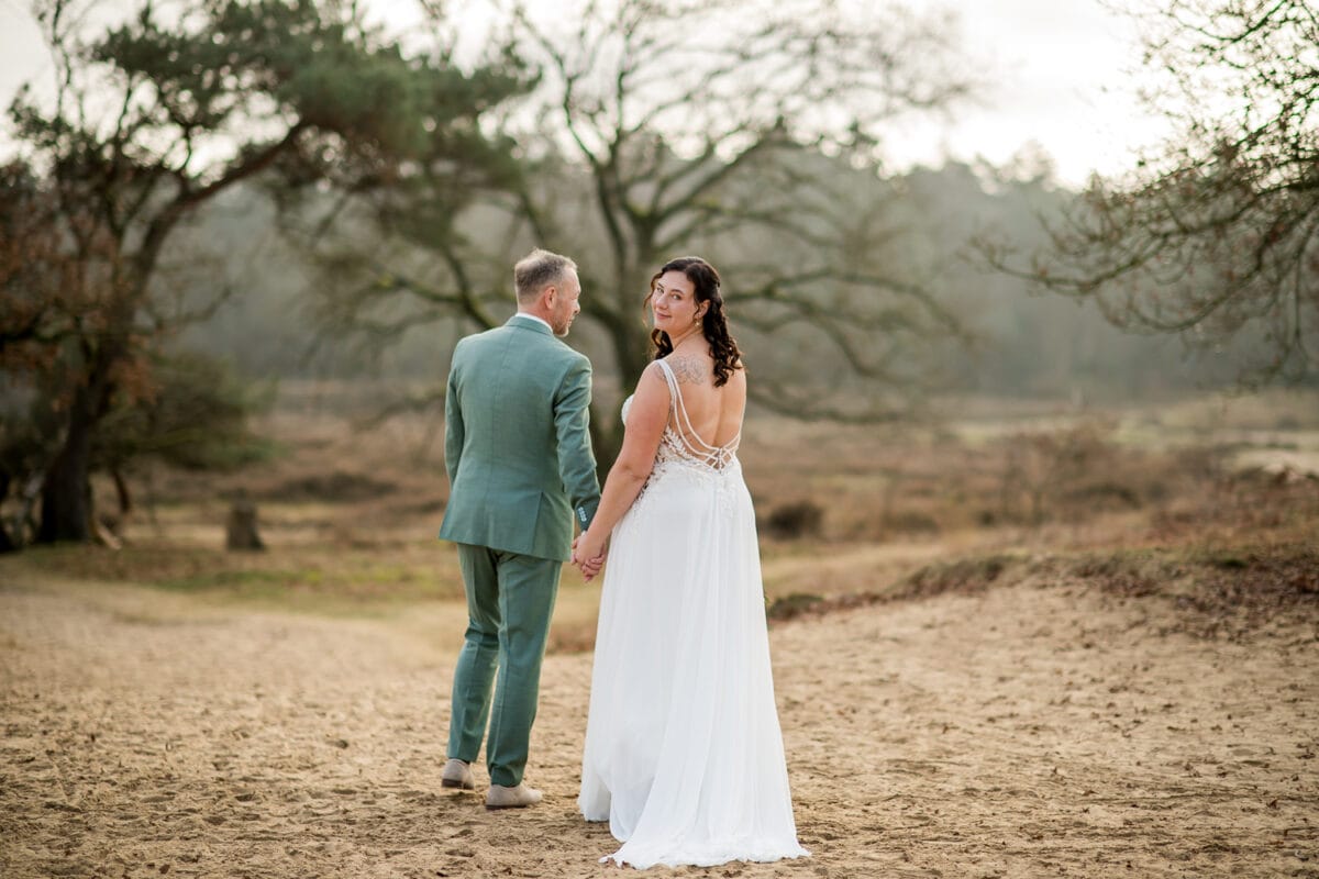 Bruidspaar hand in hand op zandpad, vrouw in witte trouwjurk kijkt om, man in groen pak, natuur en boerenkool bomen op achtergrond.