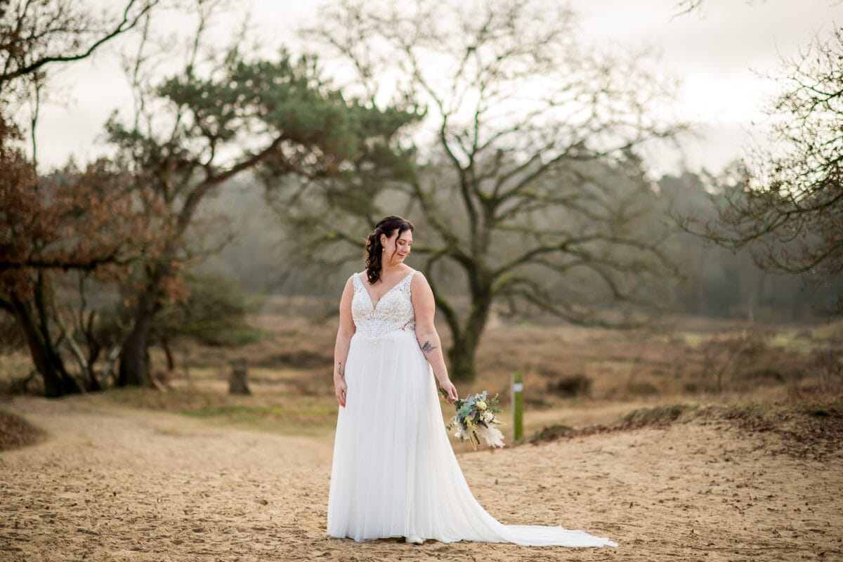Bruid in witte mouwloze trouwjurk buiten op zandgrond, boeket vasthoudend, glimlachend met bomen en bewolkte lucht op achtergrond.