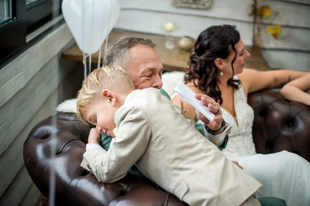 Gezin op leren bank, man in pak knuffelt blonde jongen in beige kostuum, vrouw in witte bruidsjurk, ballonnen en rustieke decoratie.
