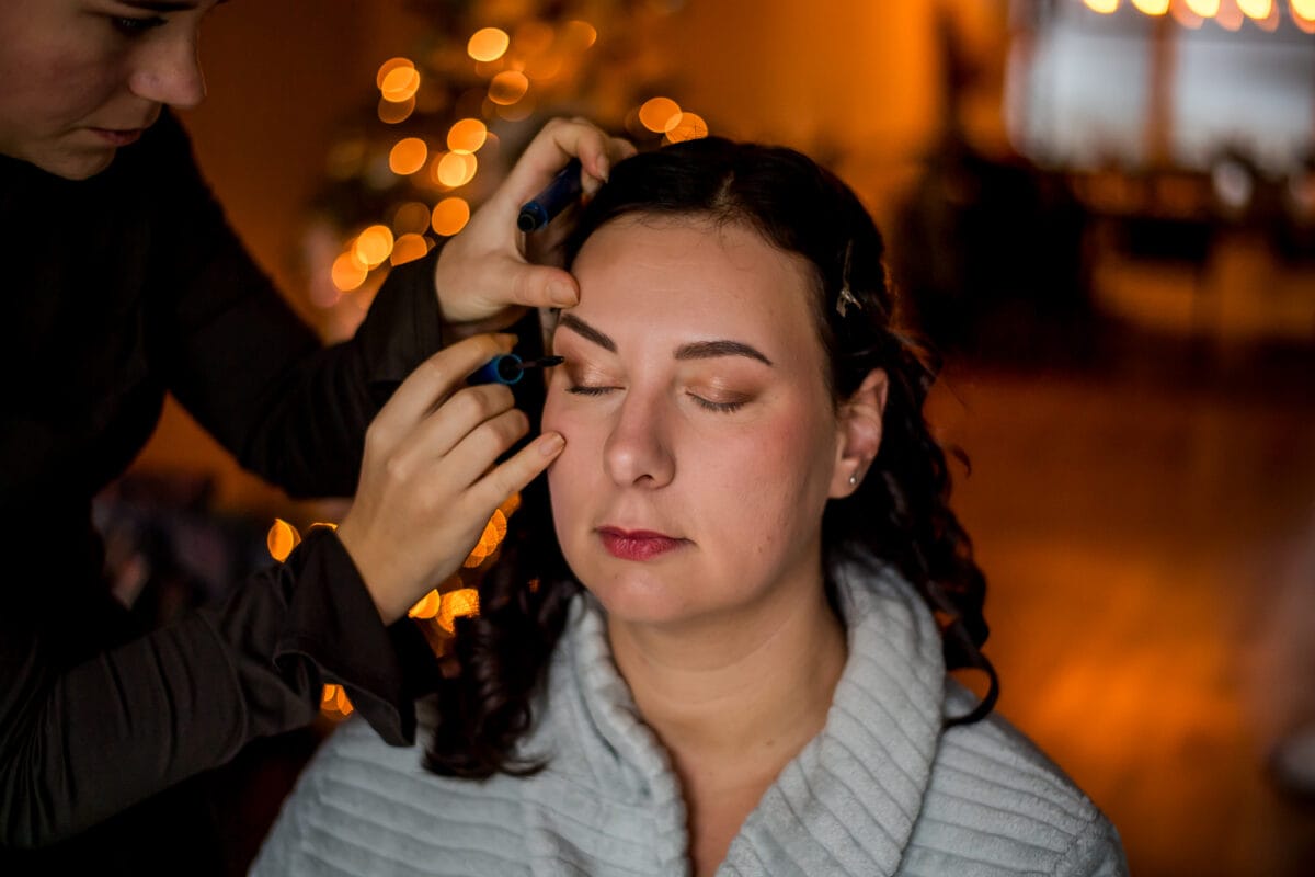 Make-up artist brengt wenkbrauwmake-up aan bij ontspannen vrouw in badjas, met warme sfeerverlichting en gouden bokeh op de achtergrond.