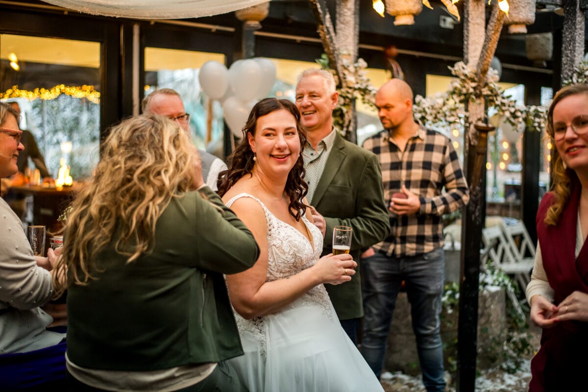 Bruid in witte jurk glimlachend, champagne vasthoudend op feestelijk binnenhuwelijksfeest met gasten, twinkelende lichtjes, ballonnen en groen.
