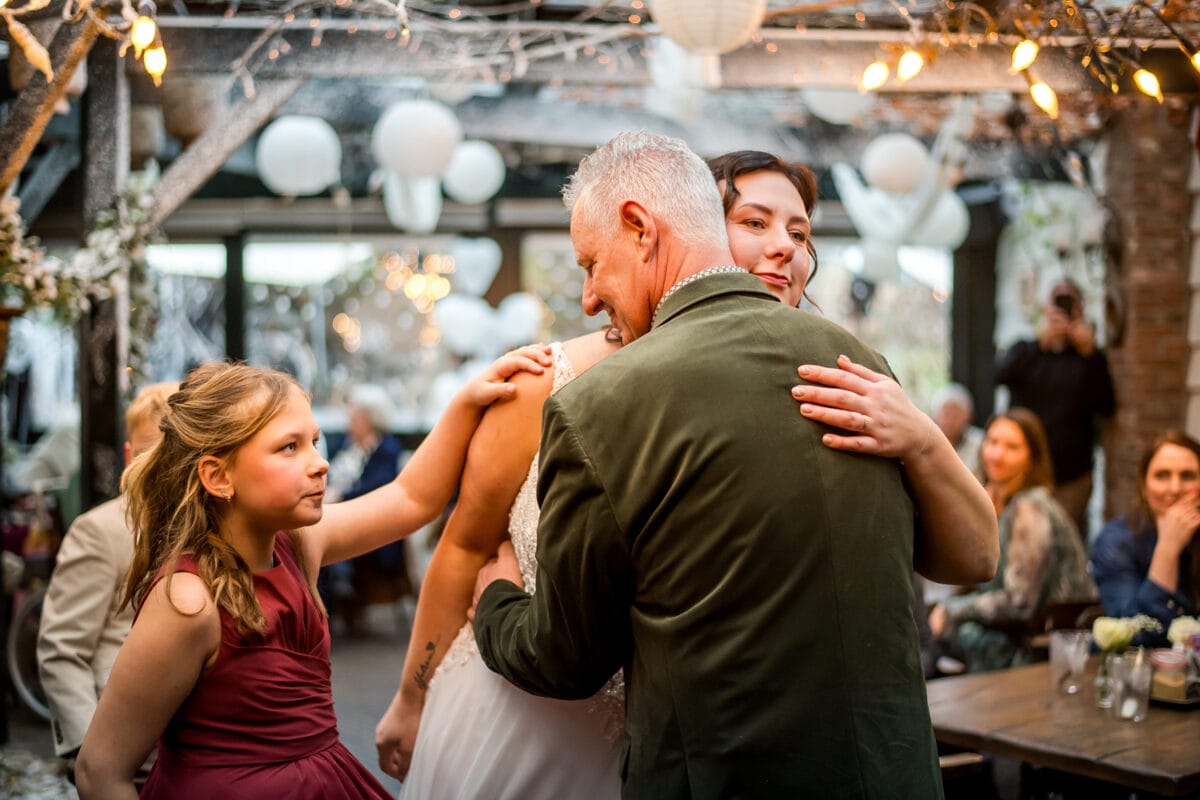 Bruiloftsfoto: bruid in witte jurk knuffelt oudere man in groen pak, meisje in bordeaux jurk raakt haar arm aan, feestelijk versierde zaal.