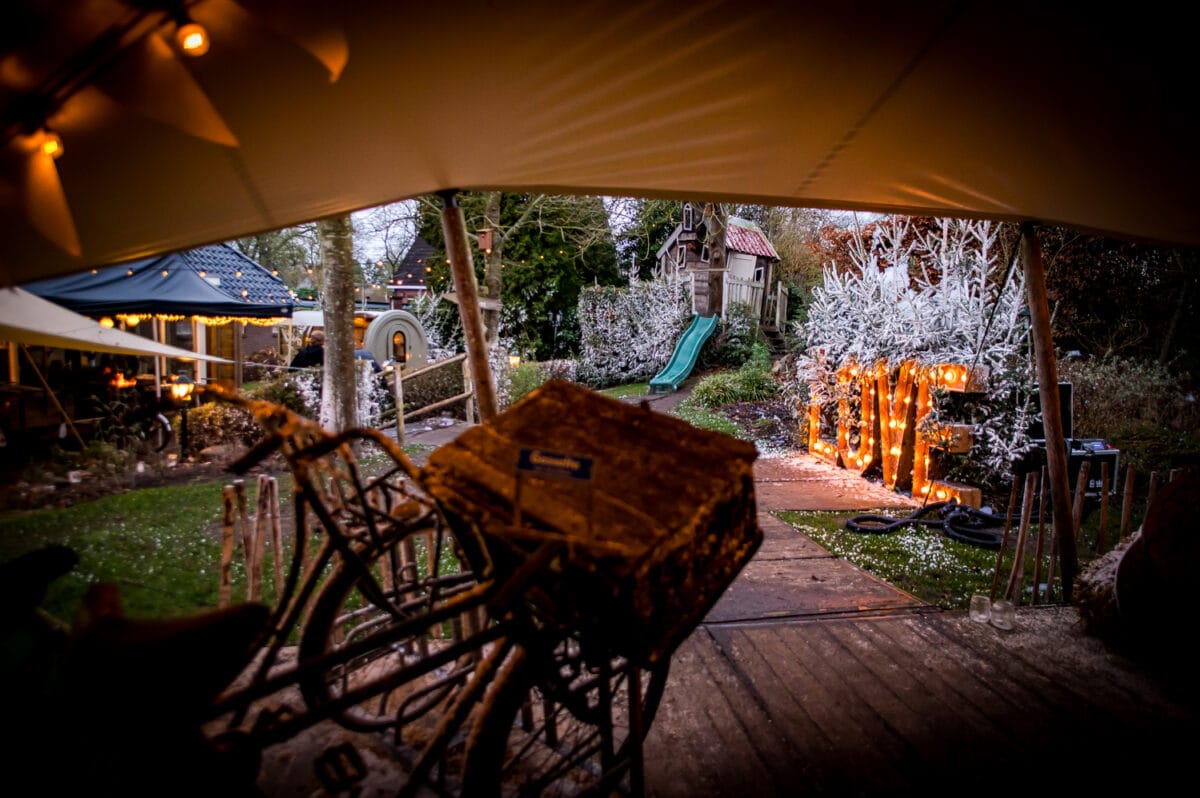 Feestelijke buitenscène in de schemering met rustieke tent, koordverlichting, oude fiets, speelglijbaan, berijpte bomen en verlicht LOVE-bord.