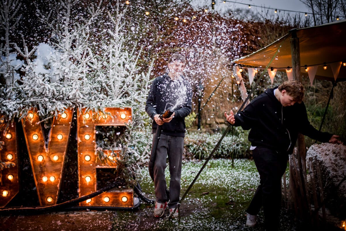 Winterpret buiten: twee mensen spelen met nepsneeuw in de buurt van gloeiend LOVE-bord, besneeuwde bomen en feestelijke verlichting. Scène van vakantieviering.