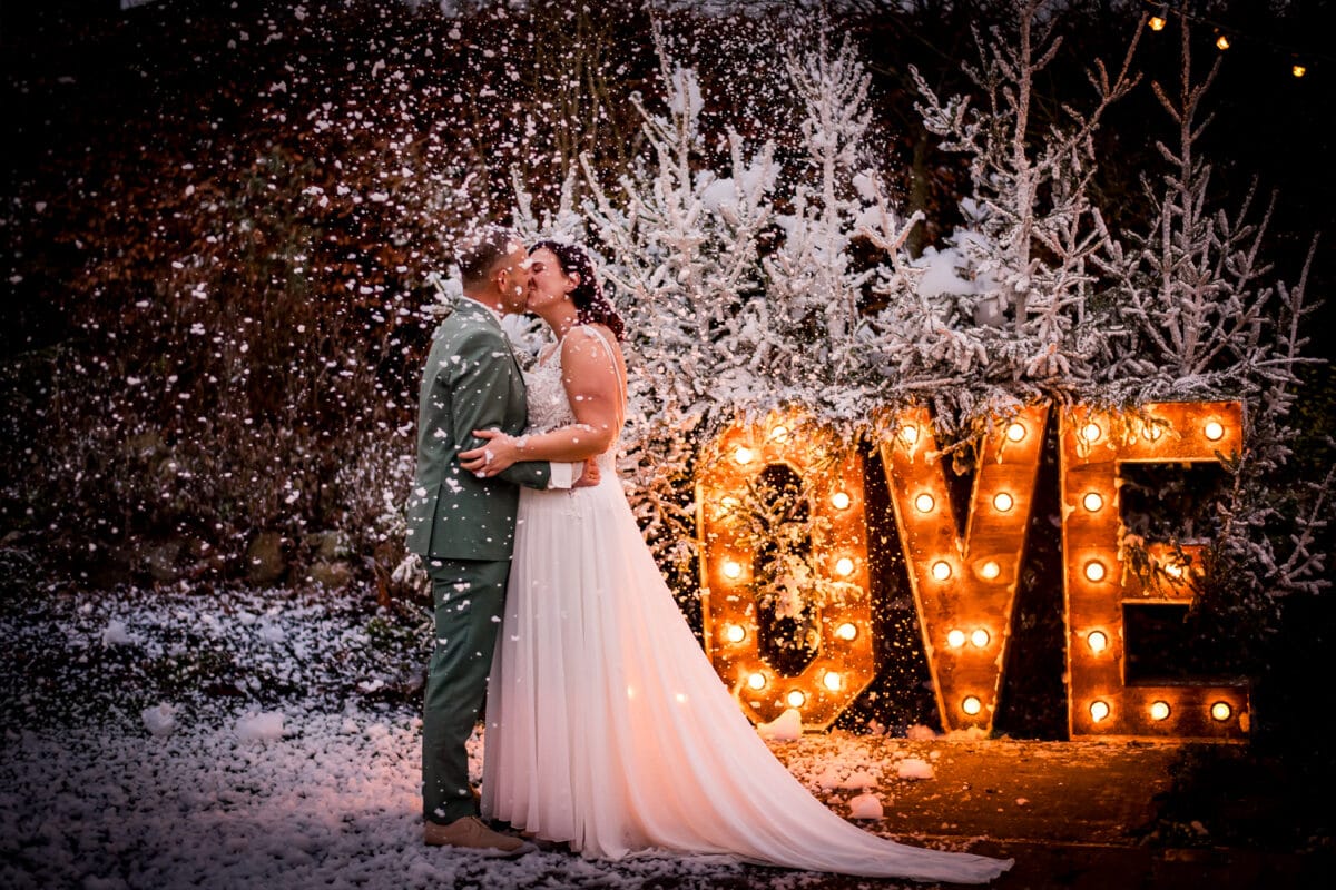 Romantische winterbruiloft: koppel kust buiten voor verlichte LOVE-letters, besneeuwde bomen en feestelijke sfeer.