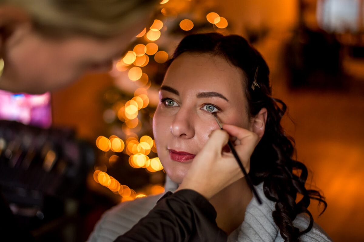Make-up artist brengt oogschaduw aan bij vrouw met donker haar, feestelijke verlichting en kerstboom op de achtergrond.