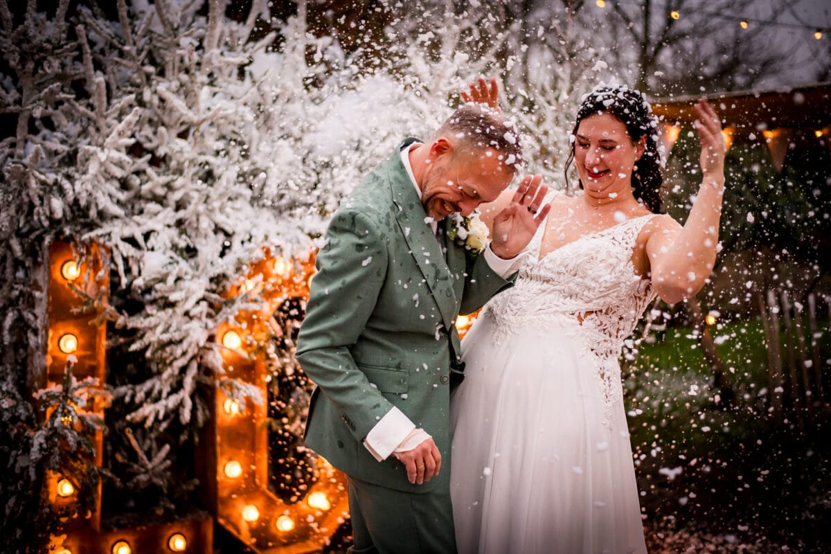 Outdoor winterbruiloft met bruid in witte jurk en bruidegom in groen pak lachend onder vallende sneeuw, feestelijke lichtjes, besneeuwde bomen.
