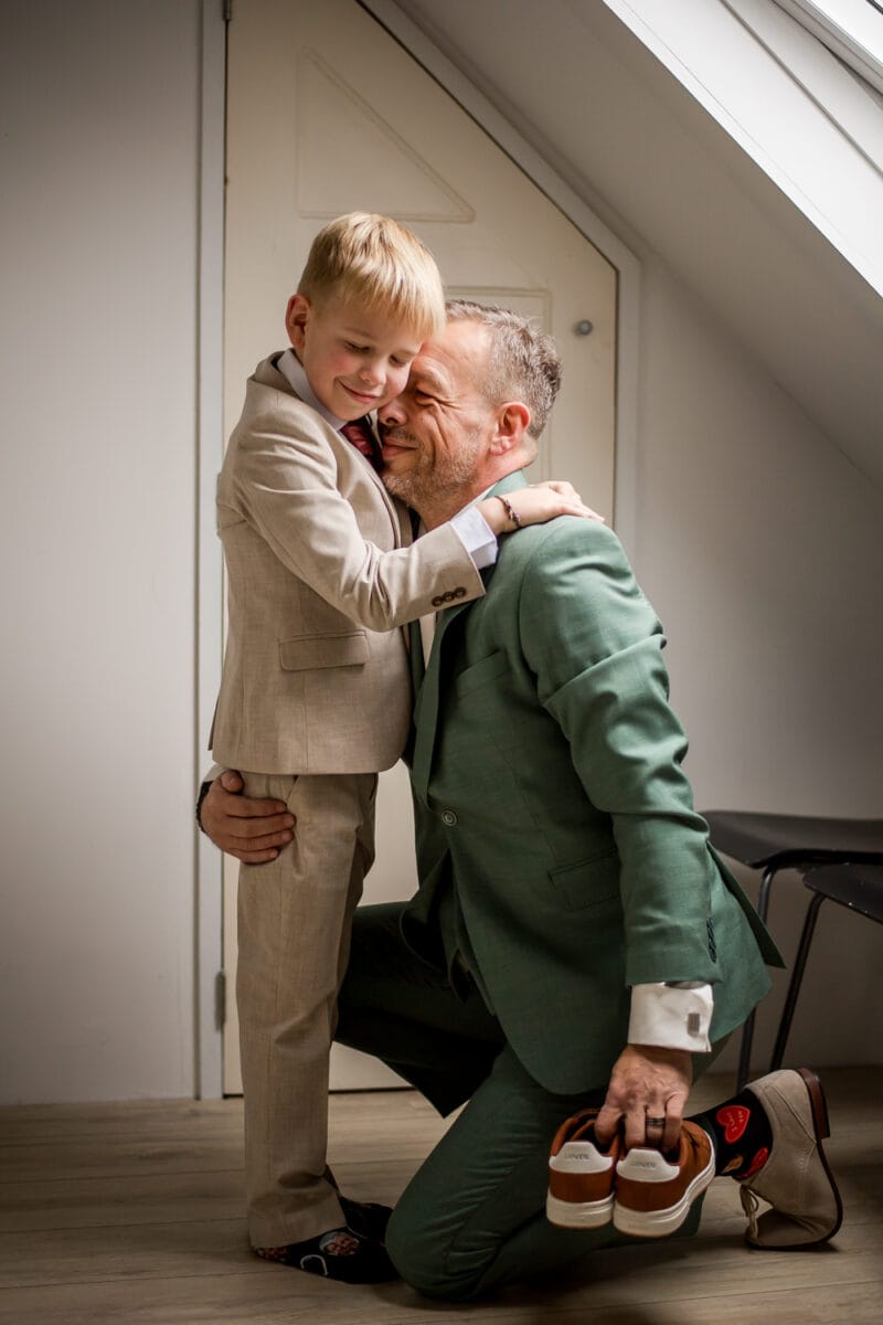 Man in groen pak omhelst lachende jongen in tan pak binnenshuis, met bruin-witte schoenen bij zonovergoten raam. Vrolijk moment van genegenheid.
