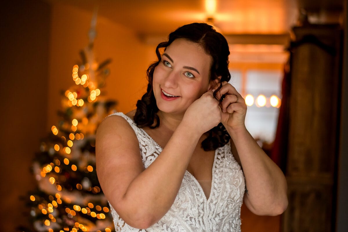 Bruid in witte kanten bruidsjurk glimlacht en doet oorbellen in huis in de buurt van een versierde kerstboom voor een feestelijke trouwsfeer.